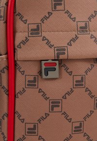 Sac à dos en tissu texturé marron avec un motif de logo Fila, des accents rouges et une plaque logo en métal avec des détails noirs et rouges.