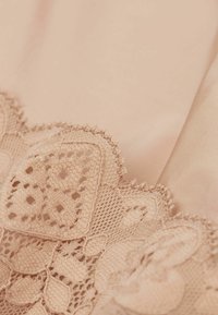Beige tyg med intrikata spetsar och blommönster, samt vågiga kanter. Slät yta med ömtålig detaljer och synliga stygn.