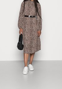 Hněd dresses s leopardím vzorem v béžové barvě, dlouhé rukávy, černý pásek a bílé tenisky. Nese černou pletenou kabelku.