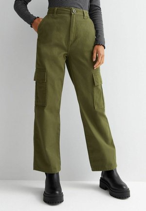 Persoon draagt olijfgroene cargobroek met klepzakken, zwarte plateauboots en een zwart-wit gestreept shirt met lange mouwen.