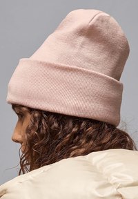 Lichtroze gebreide beanie met een omgeslagen rand, zachte textuur, gladde afwerking en een eenvoudig ontwerp, gedragen door een persoon met krullend haar.