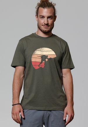 Man with beard and tied hair wears olive green t-shirt featuring silhouette of rock climber against a retro sunset circle.