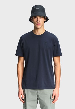 Jeune homme portant un t-shirt bleu marine, un pantalon cargo gris clair, et un chapeau seau noir avec le texte blanc « AIGLE ».