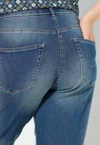 Džíny v středně modré barvě s upnutým střihem, se zadními kapsami a jemným vyblednutím, zobrazené v pase.