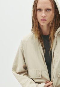 Beige Kapuzenjacke mit Reißverschluss und Druckknopfverschluss, ausgestattet mit zwei Fronttaschen und einem lässigen Schnitt, über einem dunklen Shirt getragen.
