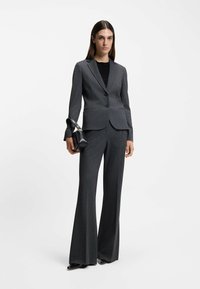 Op maat gemaakt grijs streepjespak met een getailleerde blazer en uitlopende broek. Model houdt een kleine zwarte tas vast. Soepele stof met een subtiele textuur.