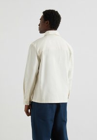 Uomo che indossa una camicia a maniche lunghe color crema e pantaloni blu scuro, in piedi con le spalle rivolte verso di noi su uno sfondo chiaro e neutro.