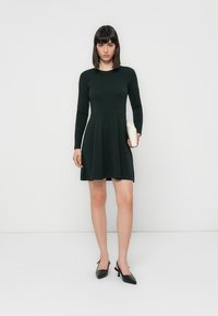 Robe longue à manches longues de couleur vert foncé, avec un corsage ajusté et une jupe évasée. Associée à des escarpins noirs à talons aiguilles et à une pochette blanche. Texture lisse.