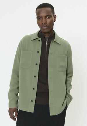 Chemise verte à boutons, fabriquée en tissu texturé, avec deux poches poitrine, un col et une fermeture éclair centrale.