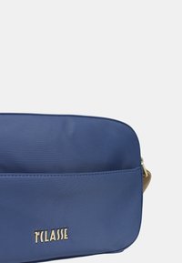 Borsa in materiale sintetico blu con una superficie liscia, forma rettangolare, dettaglio del logo in oro e una tracolla di un marrone chiaro a contrasto.