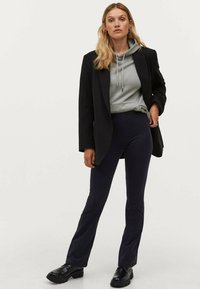 Černý blazer přes šedou mikinu, v páru s námořnickými zvonovými kalhotami a černými robustními botami. Jednoduchý střih, hladká tkanina a minimální akcenty.