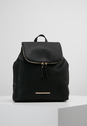 Sac à dos en cuir noir avec rabat et fermeture éclair dorée, avec deux tirettes et une petite plaque de marque rectangulaire dorée.