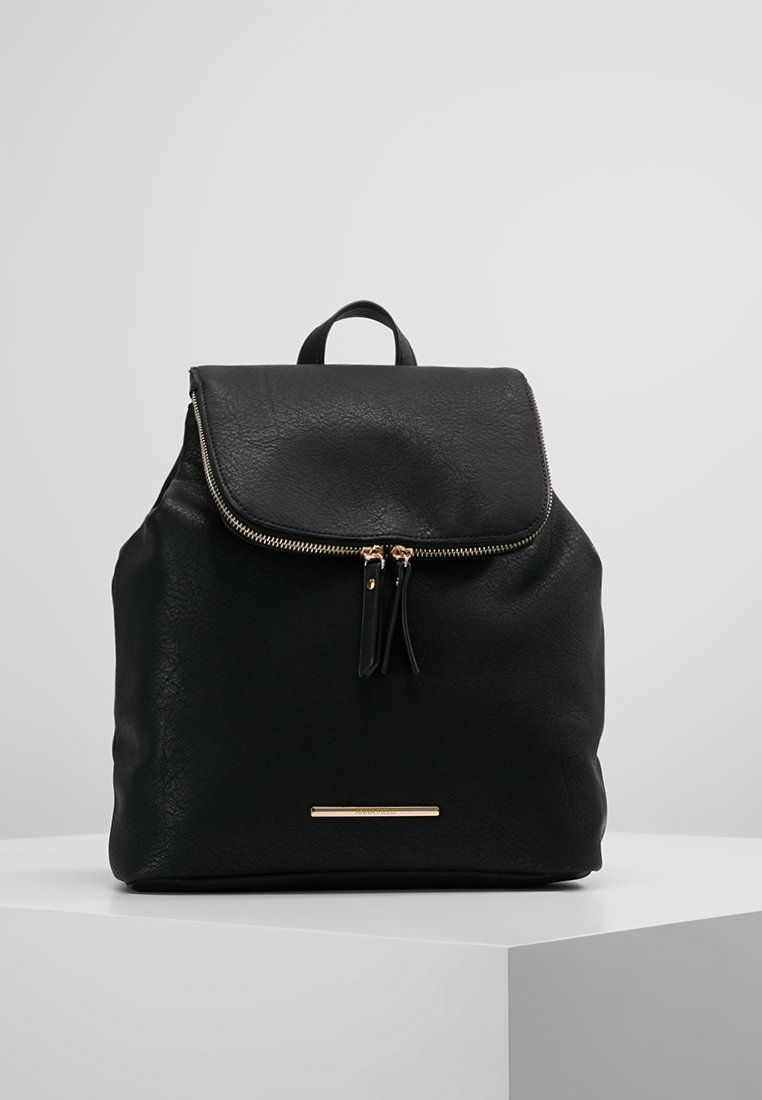 Sac à dos en cuir noir avec rabat et fermeture éclair dorée, avec deux tirettes et une petite plaque de marque rectangulaire dorée.