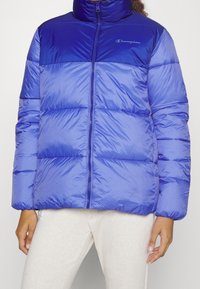 Blå vadderad jacka med hög krage, dragkedja fram och quiltade sektioner. Materialet ser glänsande ut med en slät struktur; har en logotyp på bröstet.