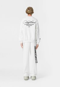 Sudadera blanca con texto negro en la parte trasera, combinada con pantalones a juego. Características notables: mangas largas, corte relajado y detalle de correa lateral.