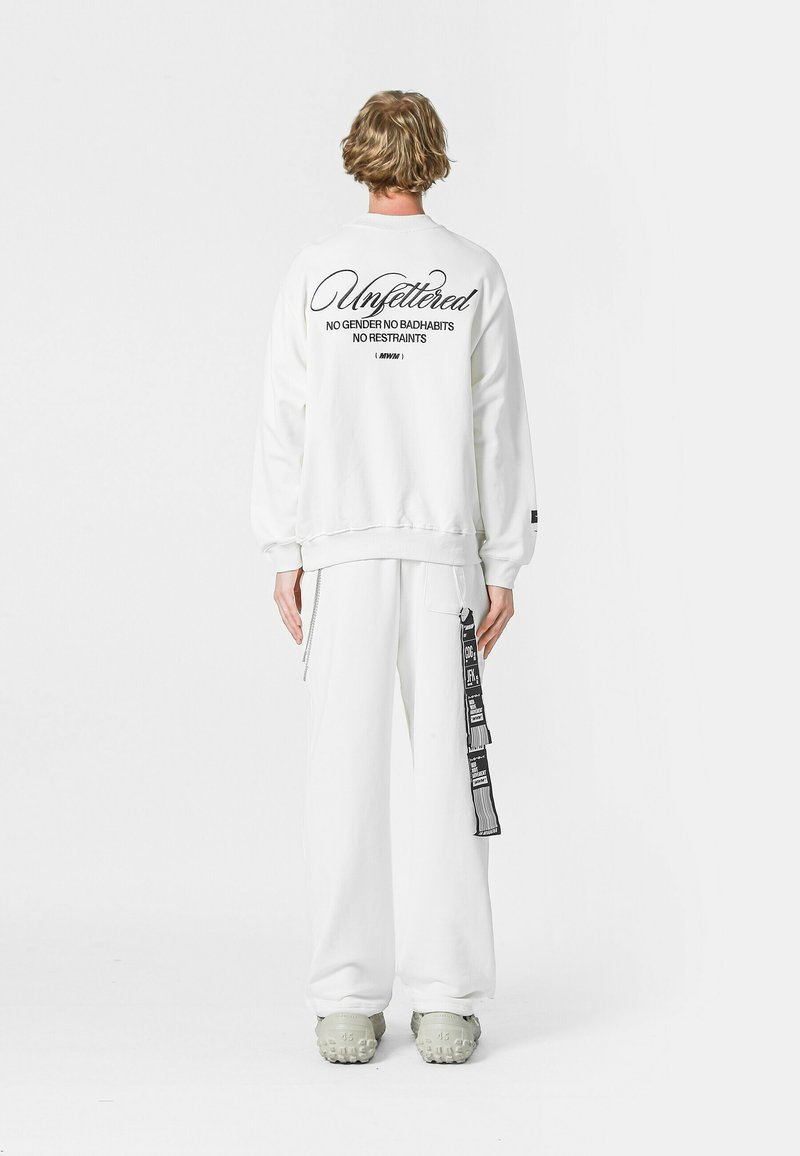 Sudadera blanca con texto negro en la parte trasera, combinada con pantalones a juego. Características notables: mangas largas, corte relajado y detalle de correa lateral.