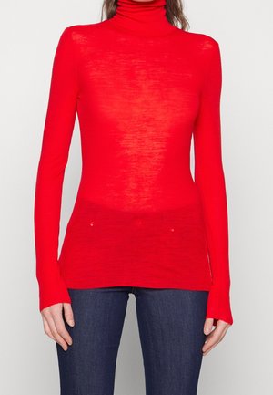 Donna con un maglione a collo alto a maniche lunghe di colore rosso acceso abbinato a jeans blu scuro, in piedi contro uno sfondo chiaro e uniforme.