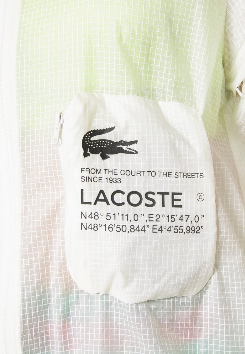 Veste légère blanche avec un motif en grille surmonté d'un vert, une poche zippée et un logo crocodile noir bien visible.