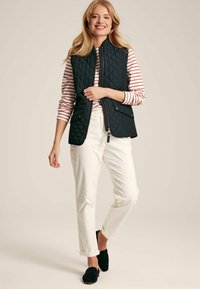 Töltött sötétkék mellény magas gallérral, cipzáros lezárással és két elülső zsebbel, csíkozott, hosszú ujjú inggel és fehér nadrággal párosítva.