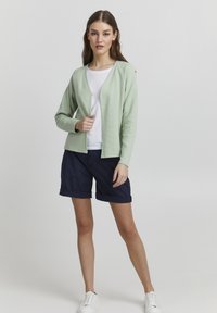 Světle zelený, žebrovaný cardigan s volným střihem a otevřeným předním designem. V páru s námořnickými zkrácenými šortkami a bílými teniskami na neutrálním pozadí.