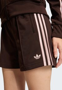 Brown athletic shorts featuring pink side stripes, elastic waistband, and embroidered logo. Smooth texture with a fitted shape.
