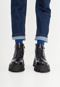 Stivali platform neri spessi con finitura lucida, design con lacci e accenti blu. Indossati con jeans di denim scuro risvoltati e calzini blu.