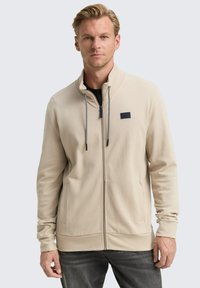 Giacca beige con zip, superficie testurizzata, cordini a contrasto e un piccolo logo nero sul petto sinistro. Presenta tasche laterali.