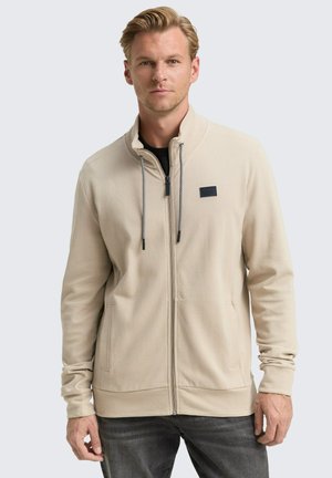 Veste zippée beige avec une surface texturée, des cordons de serrage contrastants et une petite patch logo noire sur la poitrine gauche. Elle dispose de poches latérales.