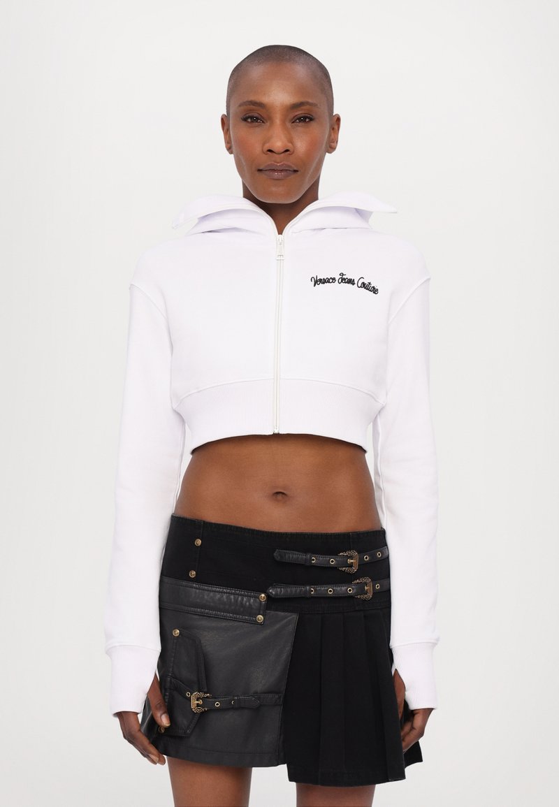 Hoodie bianco cropped con zip e ricami, abbinato a una gonna nera con dettagli in pelle, fibbie e design plissettato.