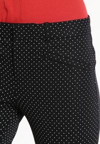 Pantalon noir avec un motif à pois blancs, comportant une poche latérale et une texture lisse. La taille se trouve au-dessus d'un haut rouge.