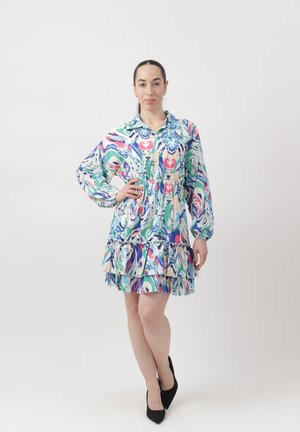 Vrouw met donker haar in een kleurrijke, met een patroon bedrukte jurk met lange mouwen, staat met één hand op haar heup en gekruiste benen, draagt zwarte hakken.