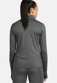 Kvinna med mörk hästsvans som bär en åtsittande långärmad grå sportjacka och matchande leggings, vänd bort från kameran mot en vit bakgrund.