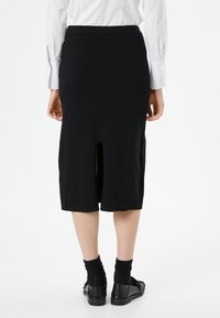 Vrouw in een zwarte kokerrok tot de knie met een split aan de achterkant, een witte lange mouwen overhemd, zwarte sokken en zwarte instapschoenen, staand met de rug naar de kijker.