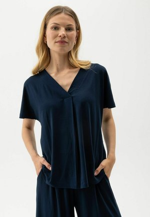 Naine sirgete blondide juustega, riietatud tumesinisesse V-kaelusega lühikeste varrukatega toppi ja sobivate laia säärega pükstega, käed taskutes.