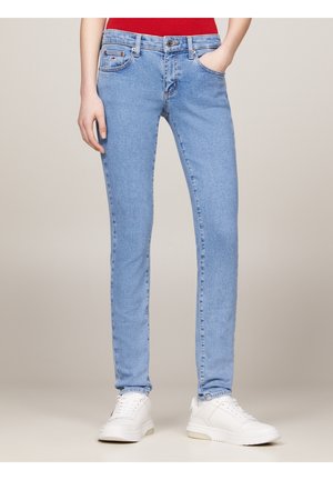 Personne portant un jean slim bleu clair, un haut rouge et des baskets blanches, debout avec une main dans la poche sur fond uni.