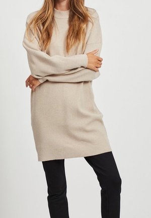 Kvinde iført en beige langærmet sweaterkjole og sorte bukser, stående med krydsede arme mod en ensfarvet hvid baggrund.