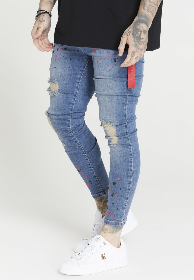 Vaqueros skinny de mezclilla en azul claro con parches desgastados en las rodillas. Presenta patrones de salpicaduras rojas y negras y una etiqueta de acento roja en el costado.