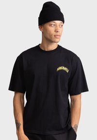 Svart bomulls T-shirt med en gul grafisk design "HAKAVELI" på vänster bröst. Modellen bär en svart ribbad mössa.