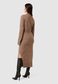 Strickkleid in Braun mit langen Ärmeln, einem Schlitz an der unteren Seite und einer taillierten Passform, kombiniert mit schwarzen kniehohen Stiefeln mit Absätzen.