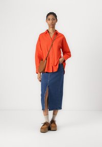Camicia rossa a maniche lunghe, gonna di jeans con spacco frontale, calze beige, scarpe con plateau marroni e una piccola borsa a tracolla marrone.