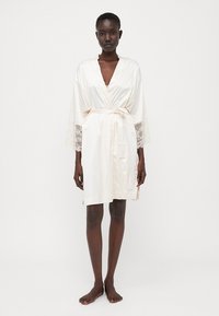 Accappatoio in seta color crema con pizzo alle maniche, dotato di una cintura in vita, di lunghezza corta e con texture morbida. Design minimalista.