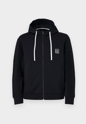 Black hoodie with zip, white drawstrings, textured black sleeves, and a small logo patch on the chest. Soft fabric with a fitted style.