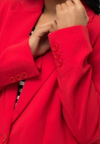 Personne en train de boutonner la manchette d'un blazer rouge vif à trois boutons rouges, portant un chemisier rayé noir et blanc en dessous.