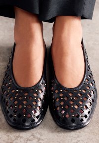 Zapatos planos negros con un diseño trenzado, que presentan recortes cuadrados y tachuelas en tono dorado, punta redondeada y una textura suave.