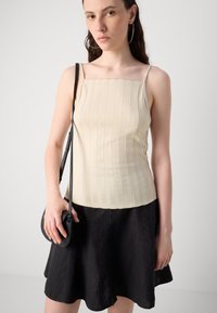 Débardeur beige à côtes avec des fines bretelles, associé à une jupe noire évasée. L'accessoire comprend un petit sac bandoulière noir.