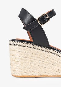 Sandalia de cuña de cuero negro con tacón de yute tipo espadrille. Presenta una hebilla en la correa del tobillo y una superficie texturizada en el tacón.