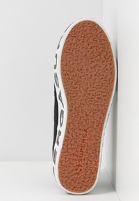 Black sneaker with textured rubber sole, featuring a pronounced orange grip pattern, white accents, and black textile upper with branding.