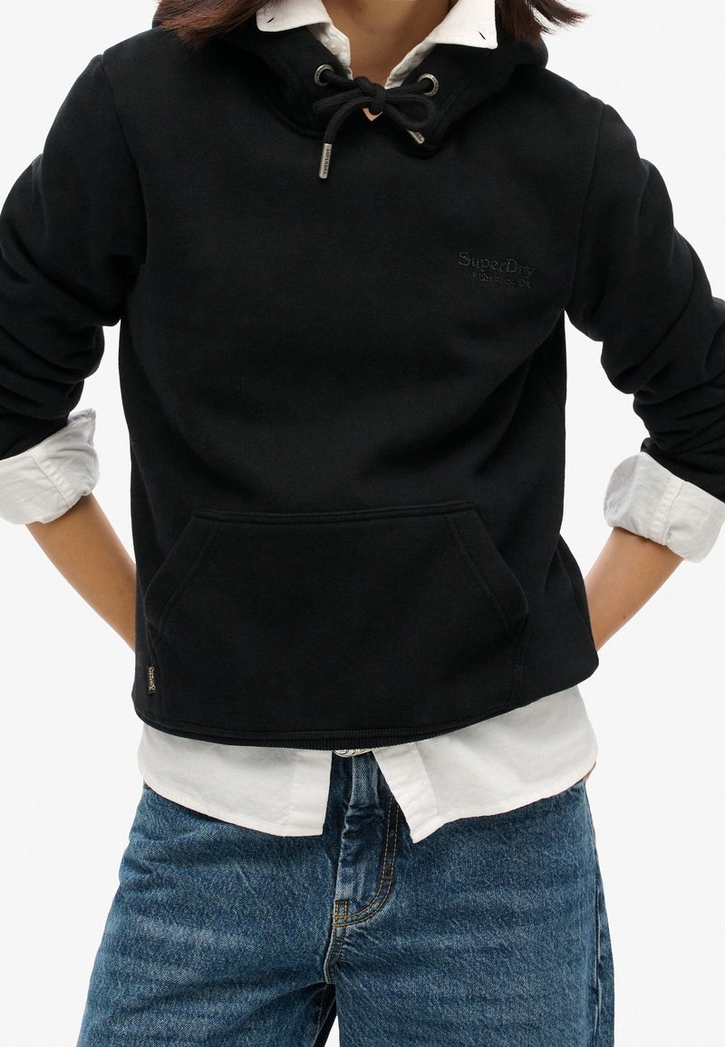 Personne portant un sweat à capuche noir par-dessus une chemise blanche boutonnée aux manches retroussées et un jean bleu, les mains posées sur les hanches.