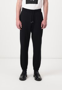 Black jogger pants with elastic waistband and cuffs. Made from soft fabric. Straight cut. Paired with a white t-shirt featuring a graphic print.
