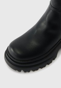 Černá kožená bota s texturovaným povrchem, kulatou špičkou a silnou gumovou podrážkou s hlubokými drážkami pro trakci.